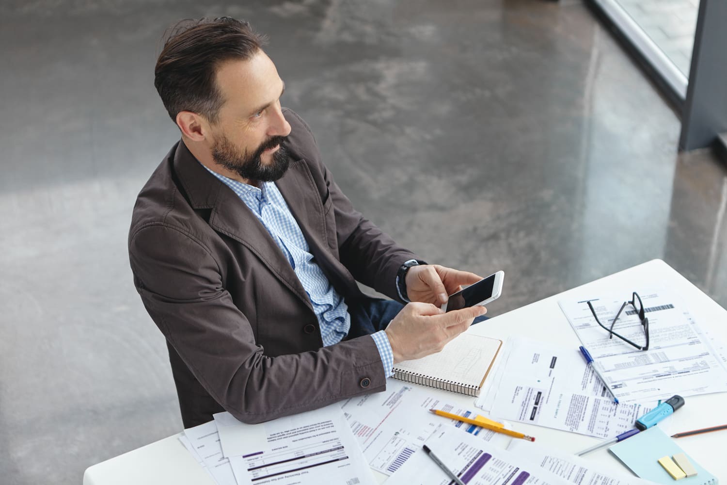 Un professionnel consulte son smartphone à son bureau, entouré de documents et de dossiers financiers, dans un environnement de travail moderne.