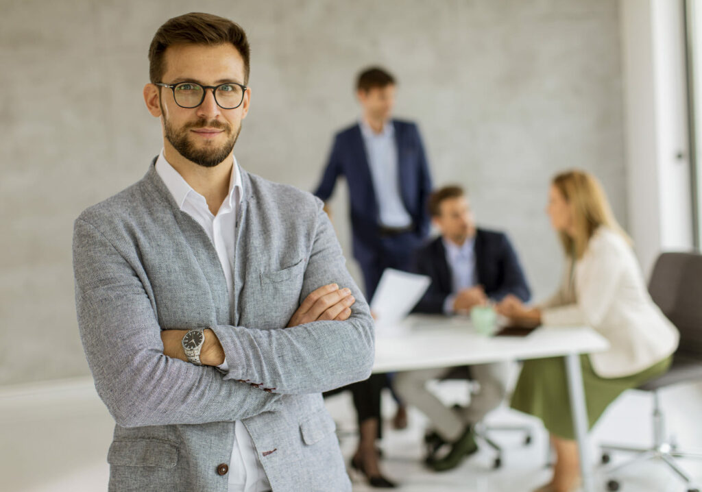 Un professionnel en costume gris et lunettes, bras croisés, se tient au premier plan d'une salle de réunion. Une équipe échange autour d'une table en arrière-plan.