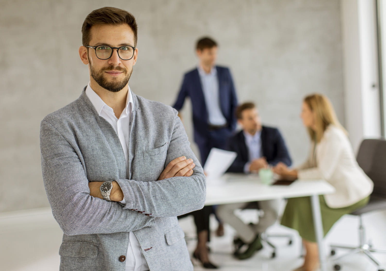 Un professionnel en costume gris et lunettes, bras croisés, se tient au premier plan d'une salle de réunion. Une équipe échange autour d'une table en arrière-plan.