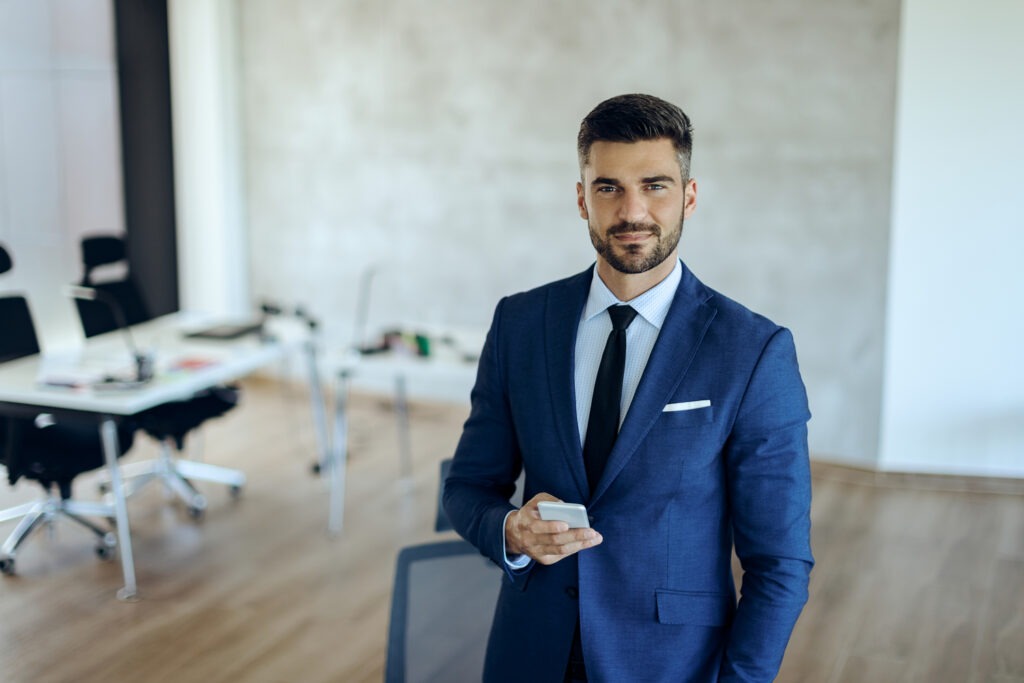 Homme en costume dans un environnement professionnel, tenant un smartphone et regardant l’objectif