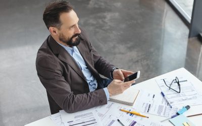 Un professionnel consulte son smartphone à son bureau, entouré de documents et de dossiers financiers, dans un environnement de travail moderne.