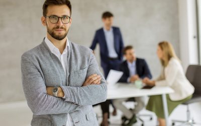 Un professionnel en costume gris et lunettes, bras croisés, se tient au premier plan d'une salle de réunion. Une équipe échange autour d'une table en arrière-plan.